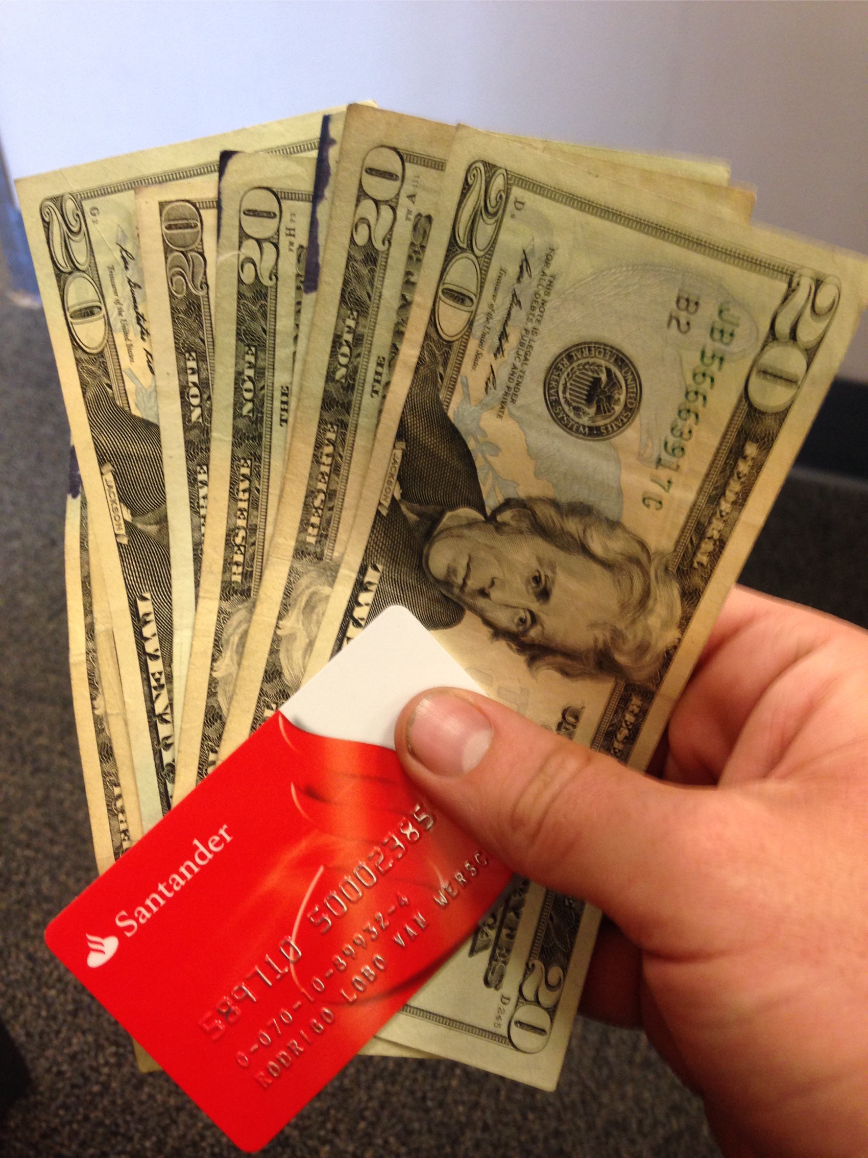 A hand holding five twenty dollar bills and a red bank card