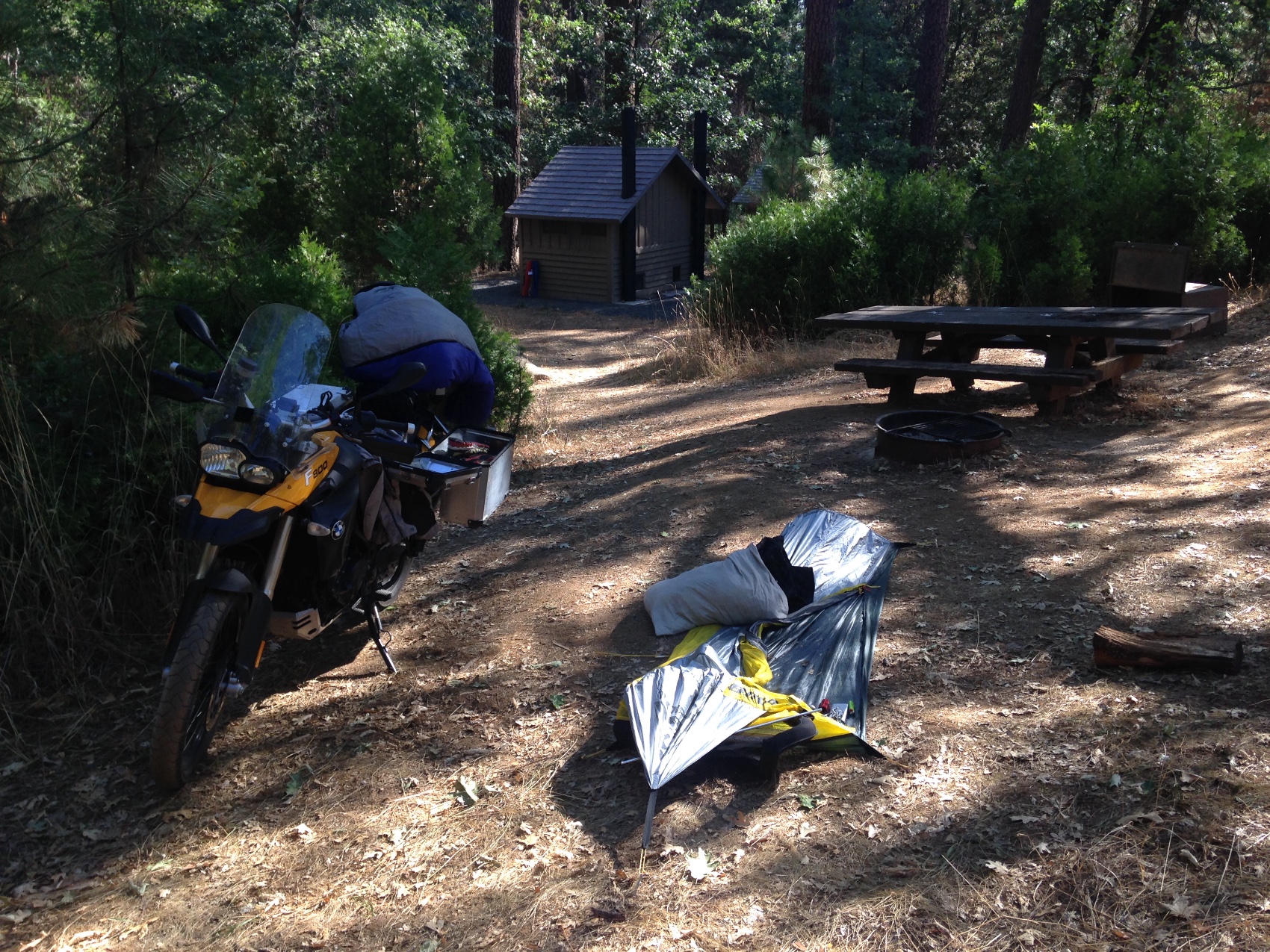 The motorcycle and a tent beside a forest campsite table and fire ring