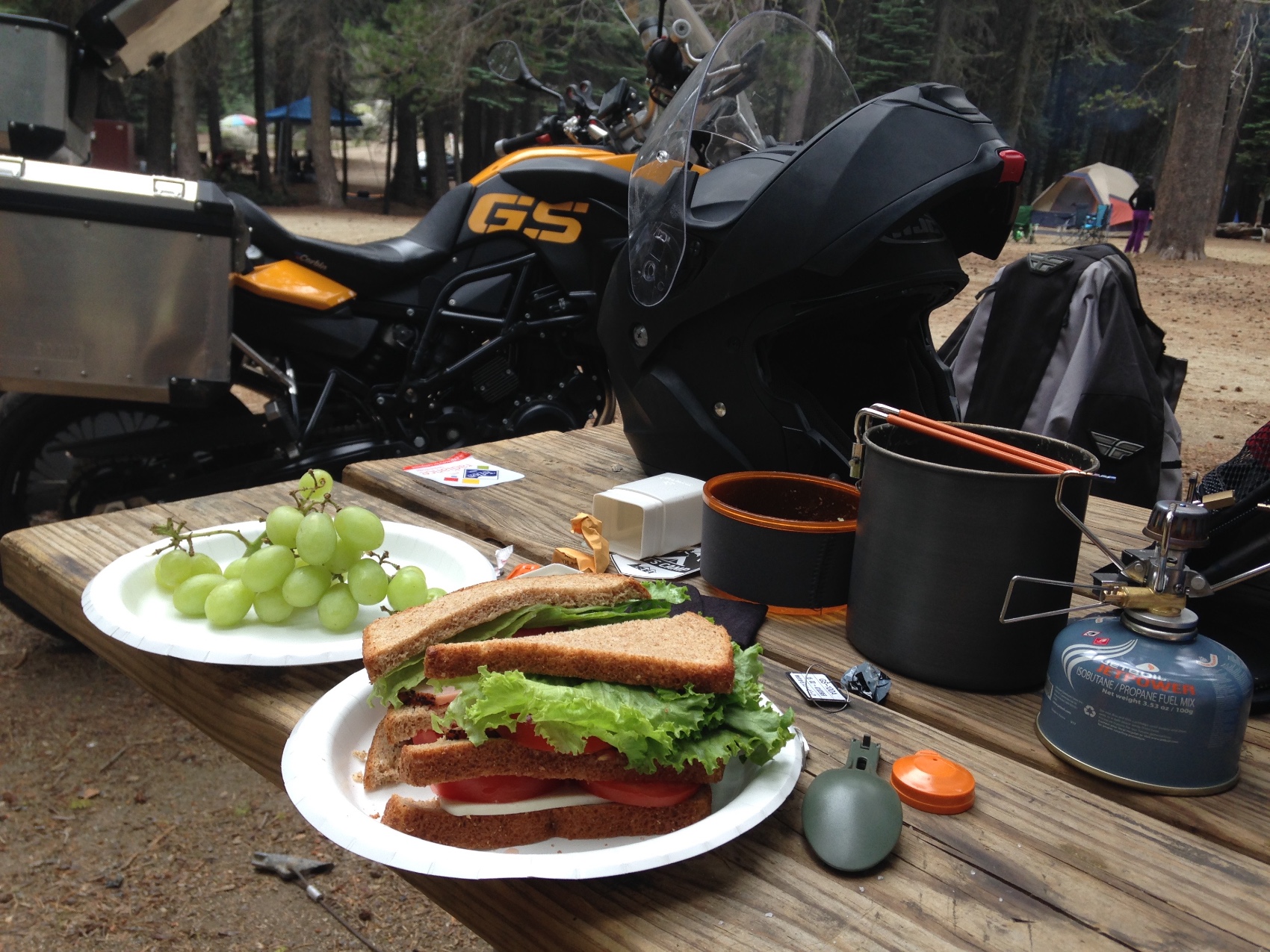 A sandwich, grapes and a camping stove on a picnic table beside the motorcycle