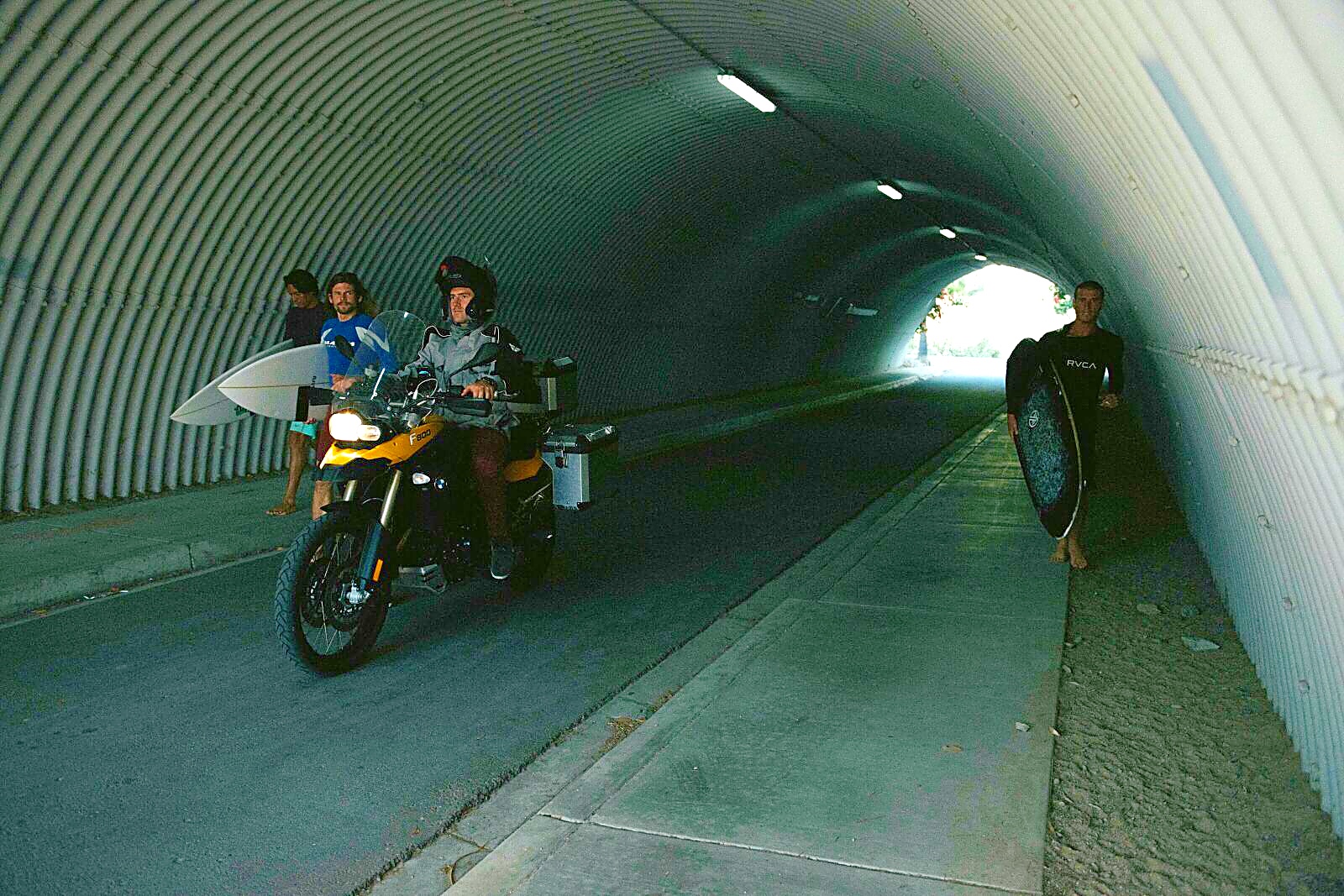 Riding through a pedestrian tunnel past surfers carrying boards