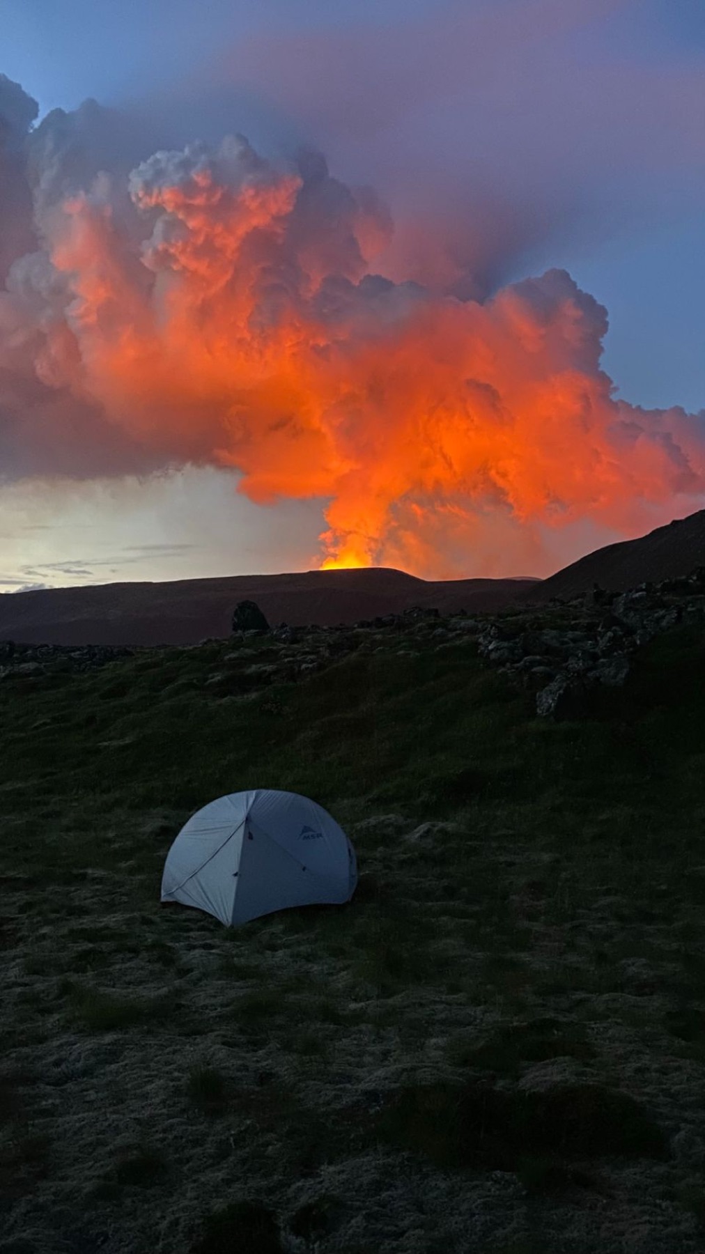 A small tent pitched on a ridge with the erupting volcano glowing behind it