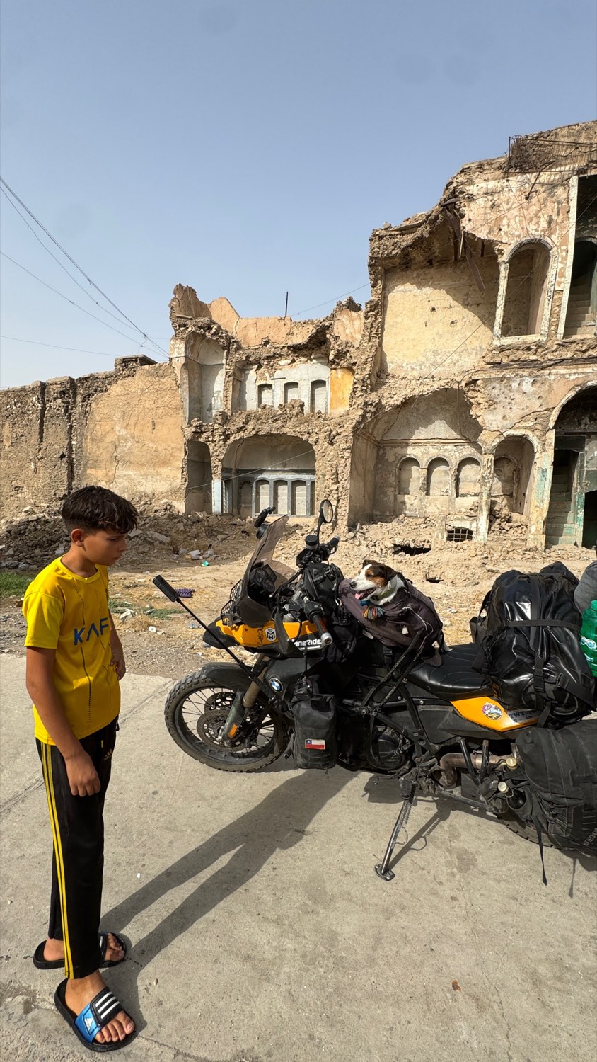 A boy in a yellow shirt looking at the loaded motorcycle in front of ruined buildings