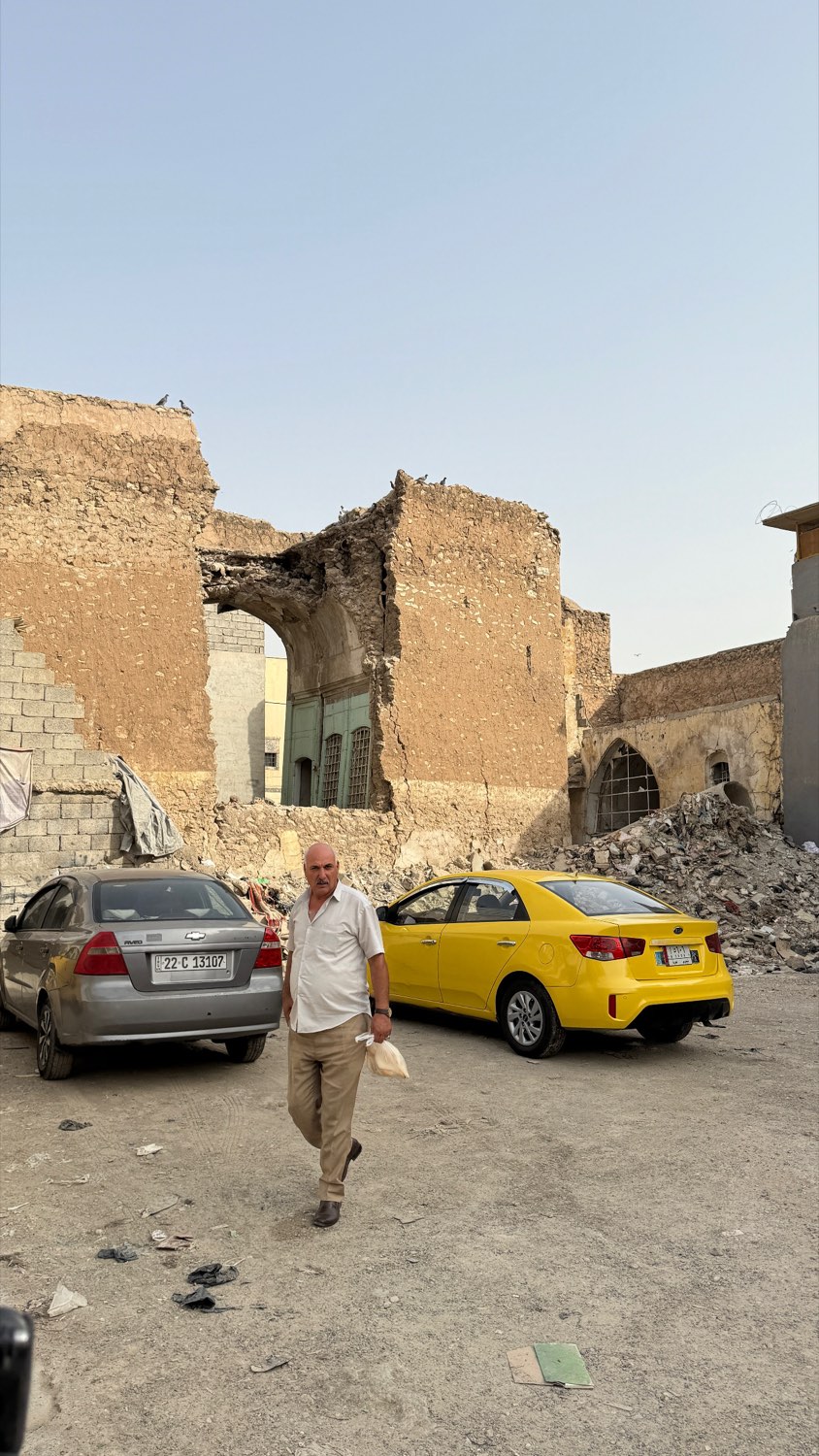 A man walking past collapsed buildings in Mosul with cars parked among the rubble