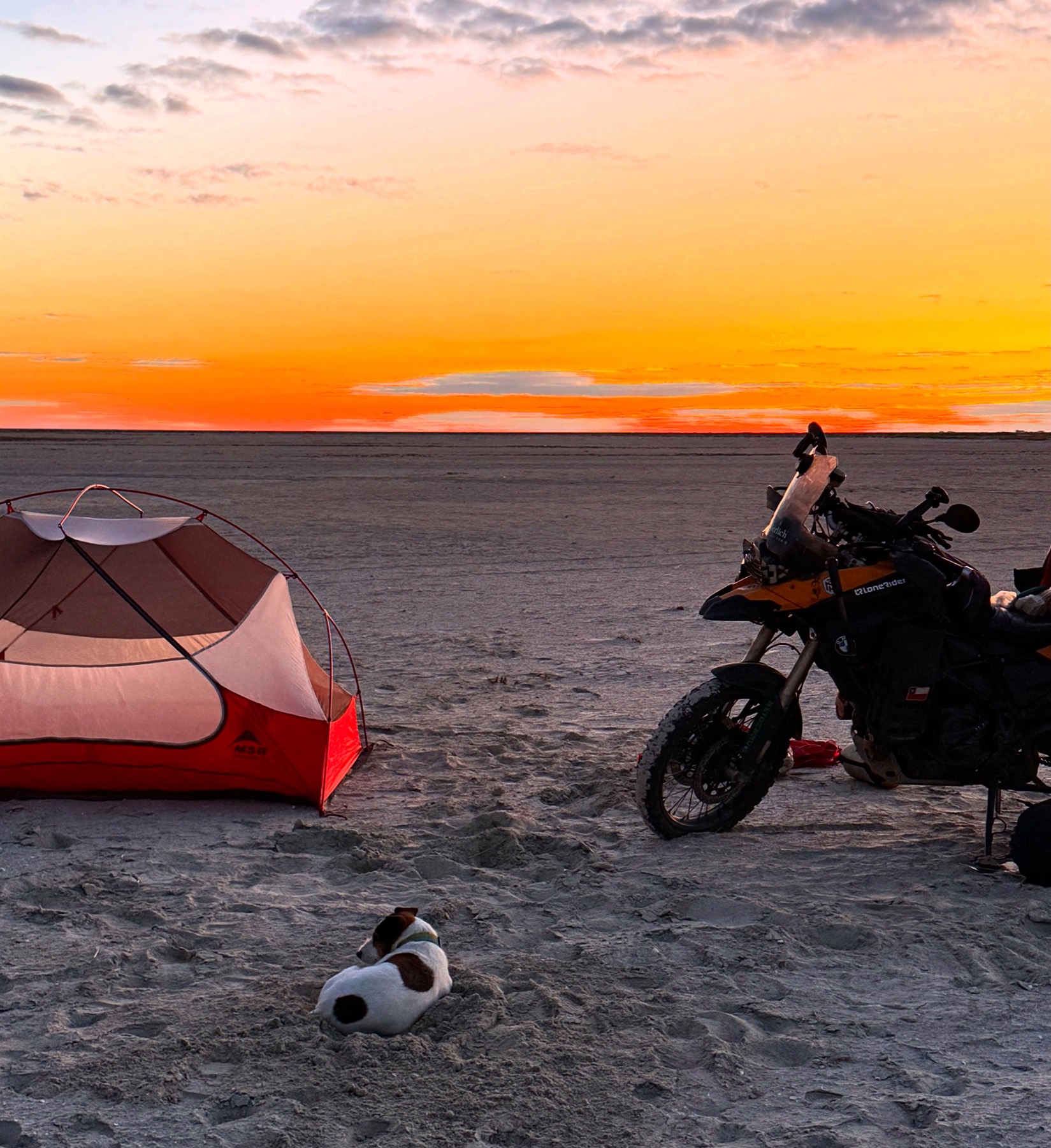 A tent and the loaded motorcycle on a salt flat at sunset, with Jacko lying in the sand