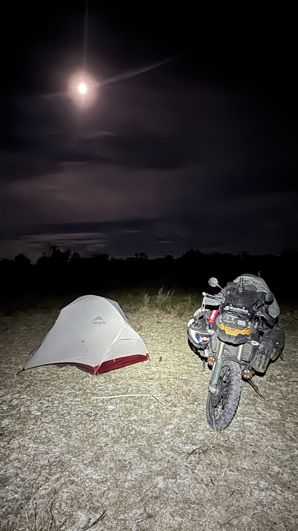 The tent and motorcycle lit under the moon on the empty steppe