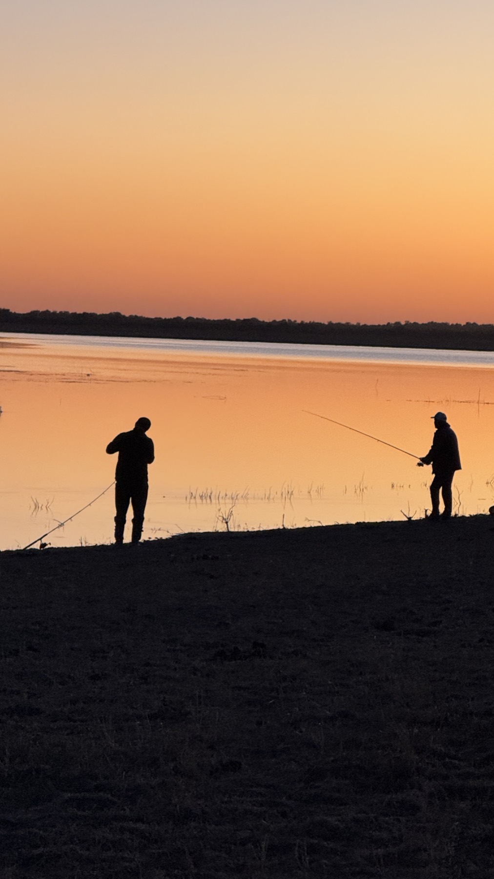 Two people fishing on a lake shore at sunset