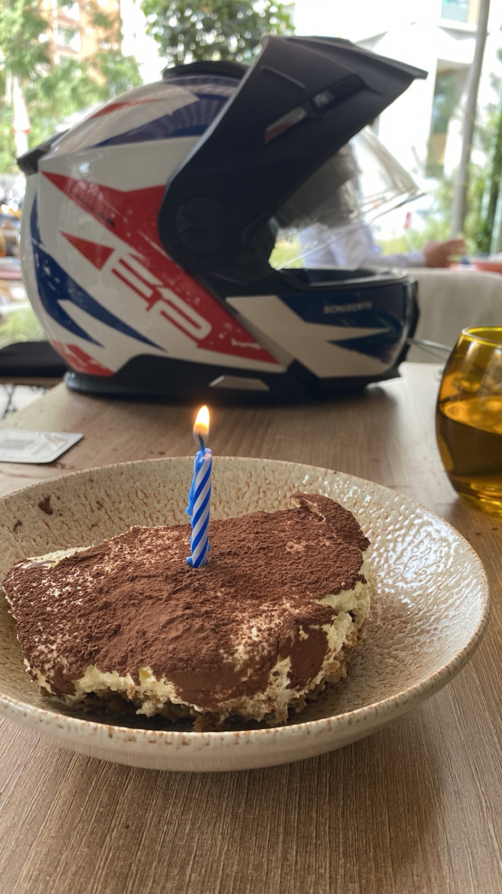 A tiramisu with a lit birthday candle on a cafe table, with a motorcycle helmet beside it