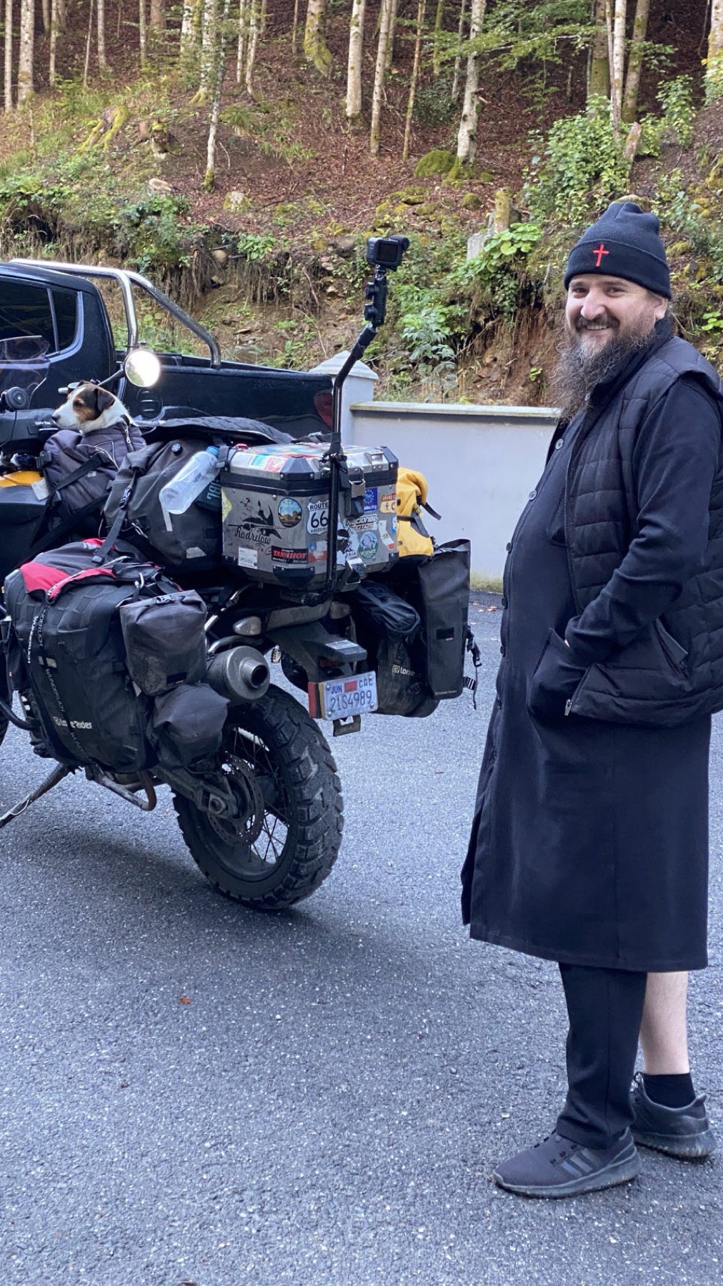 An Orthodox monk in a black cassock standing beside the loaded motorcycle