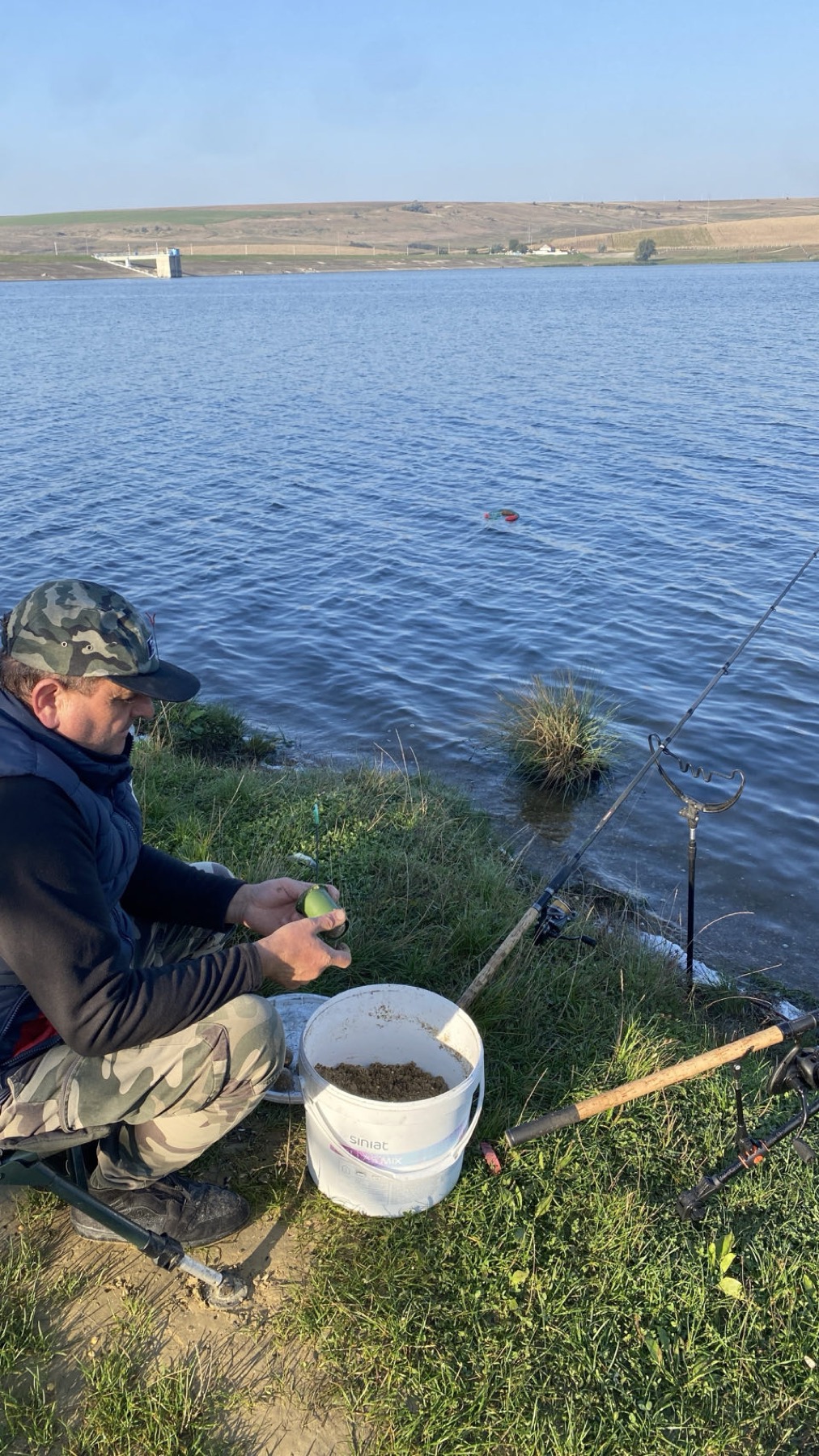 A fisherman sitting on the bank of the reservoir with rods and a bucket of bait