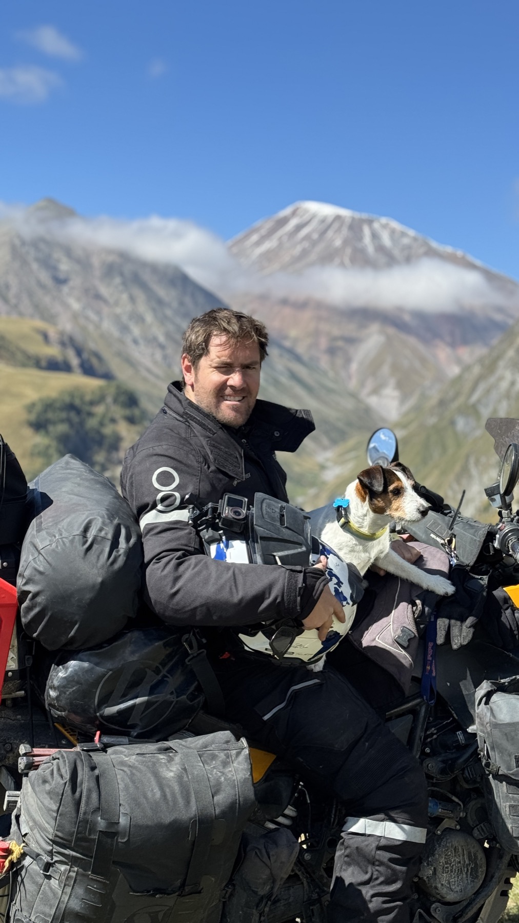 Rodrigo on the loaded motorcycle holding Jacko, with the snow cone of Mount Kazbek behind