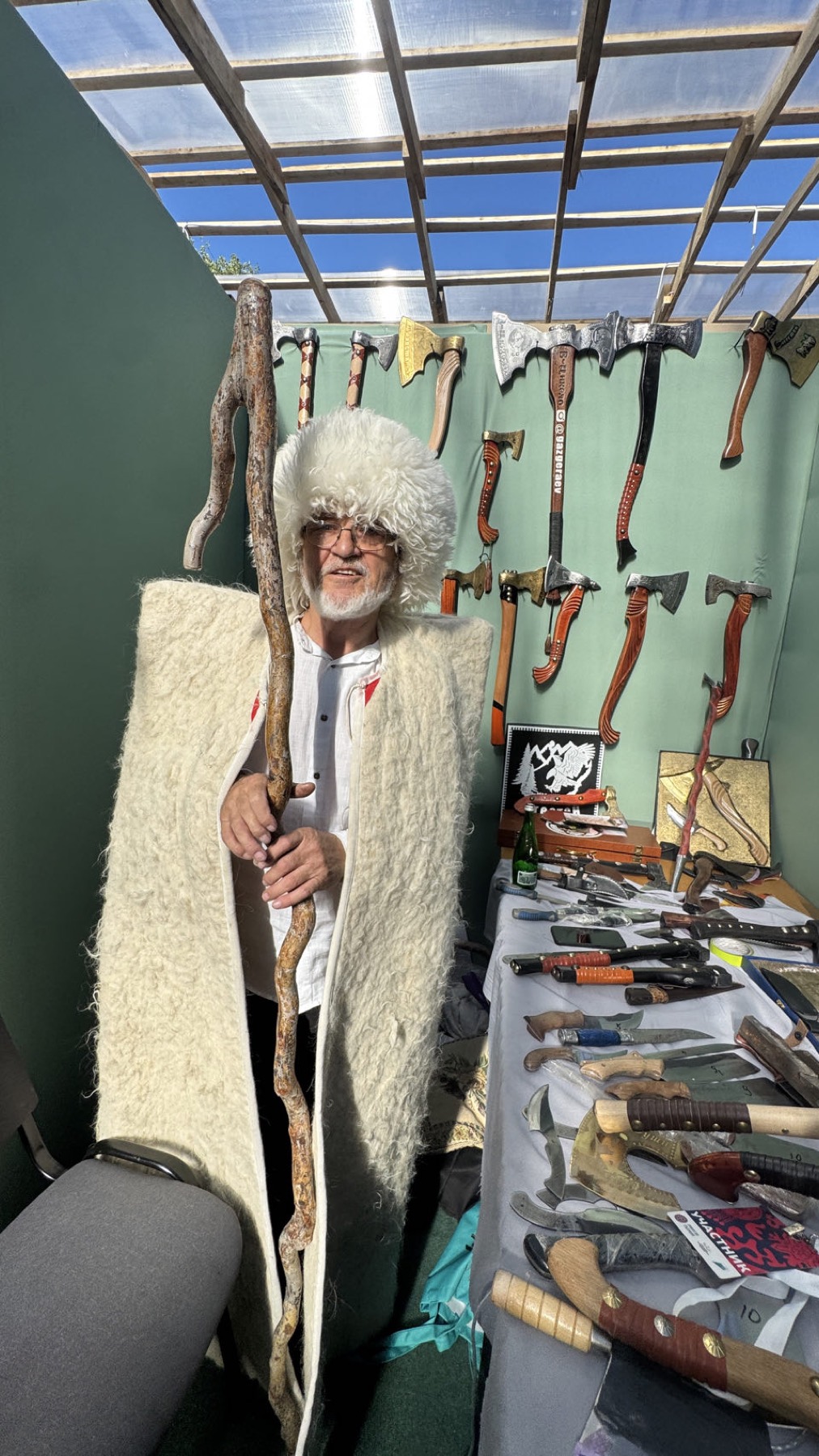 A craftsman in a sheepskin cloak beside a wall of axes and knives