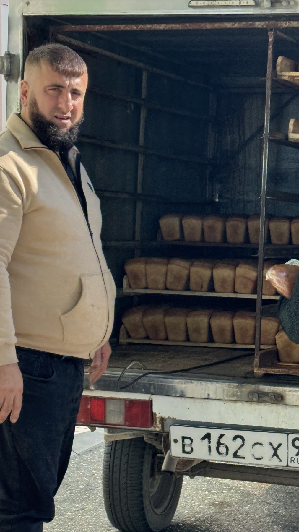 A Chechen man standing beside his delivery van, the shelves inside stacked with loaves of bread