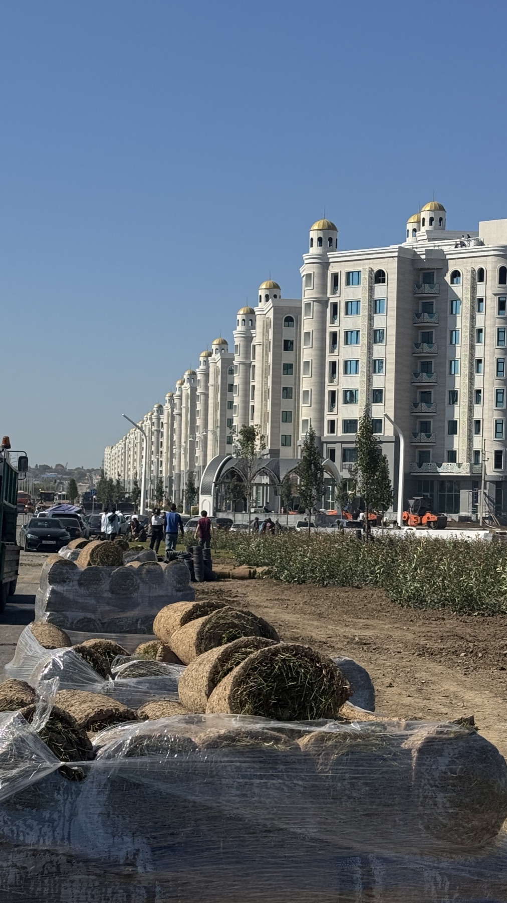 A long row of identical new white apartment blocks with golden domes, rolls of turf waiting on the ground