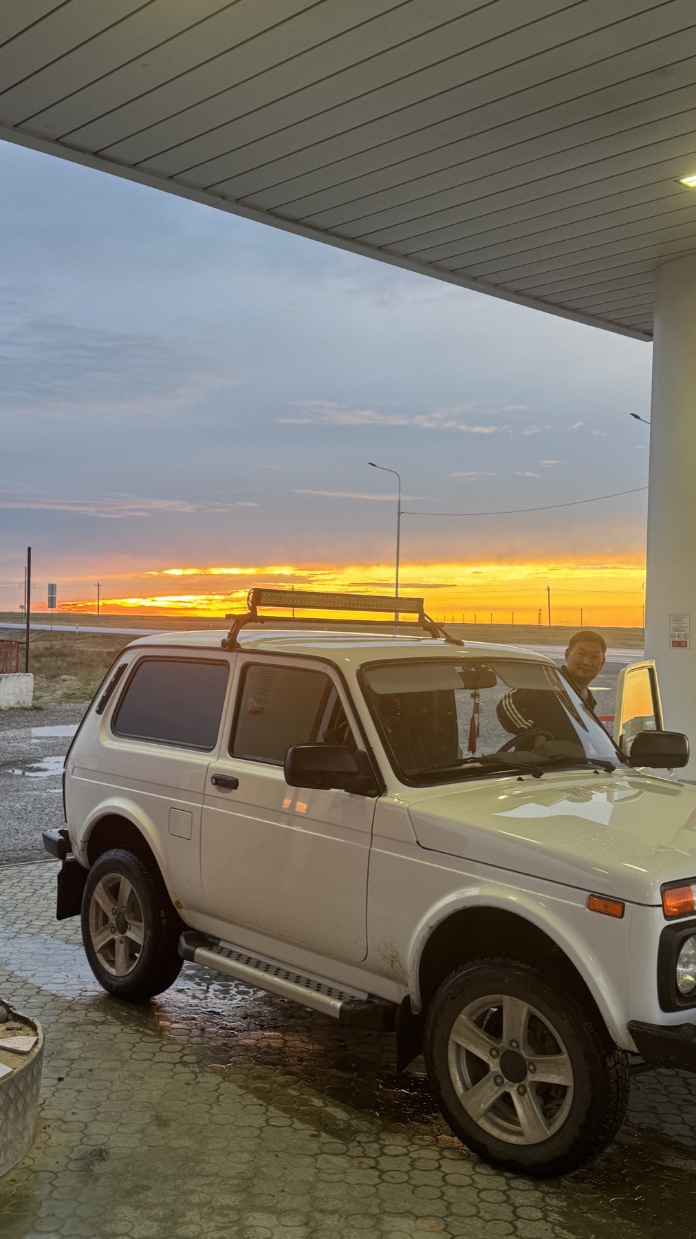 A white Lada Niva at the pumps at sunset, its driver standing by the door