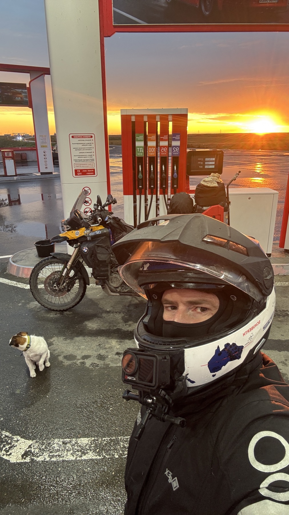 Rodrigo in his helmet at a petrol station against the sunset, with the loaded motorcycle and Jacko behind him