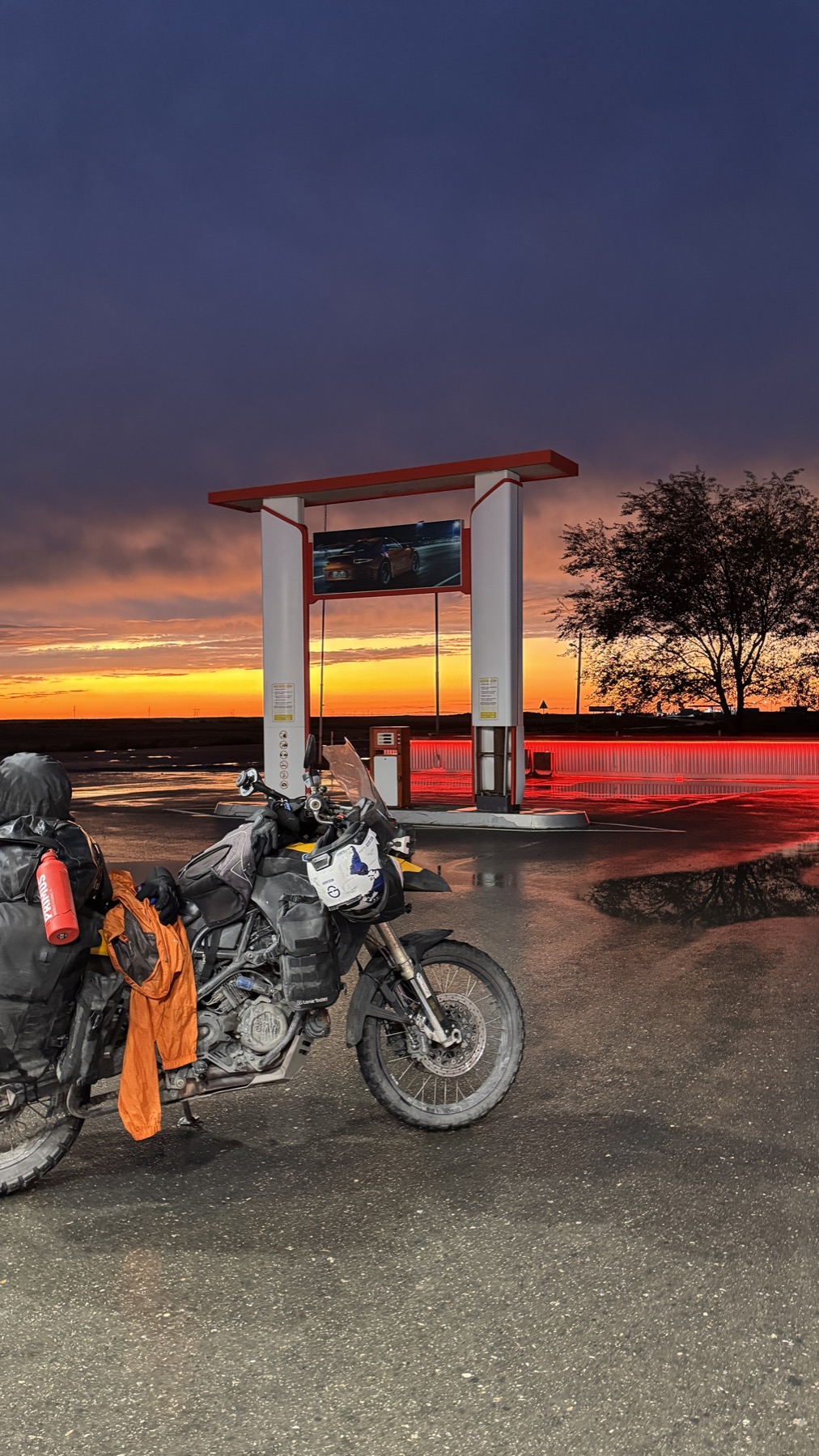 The loaded motorcycle parked at an empty petrol station on the steppe under a purple and orange sky