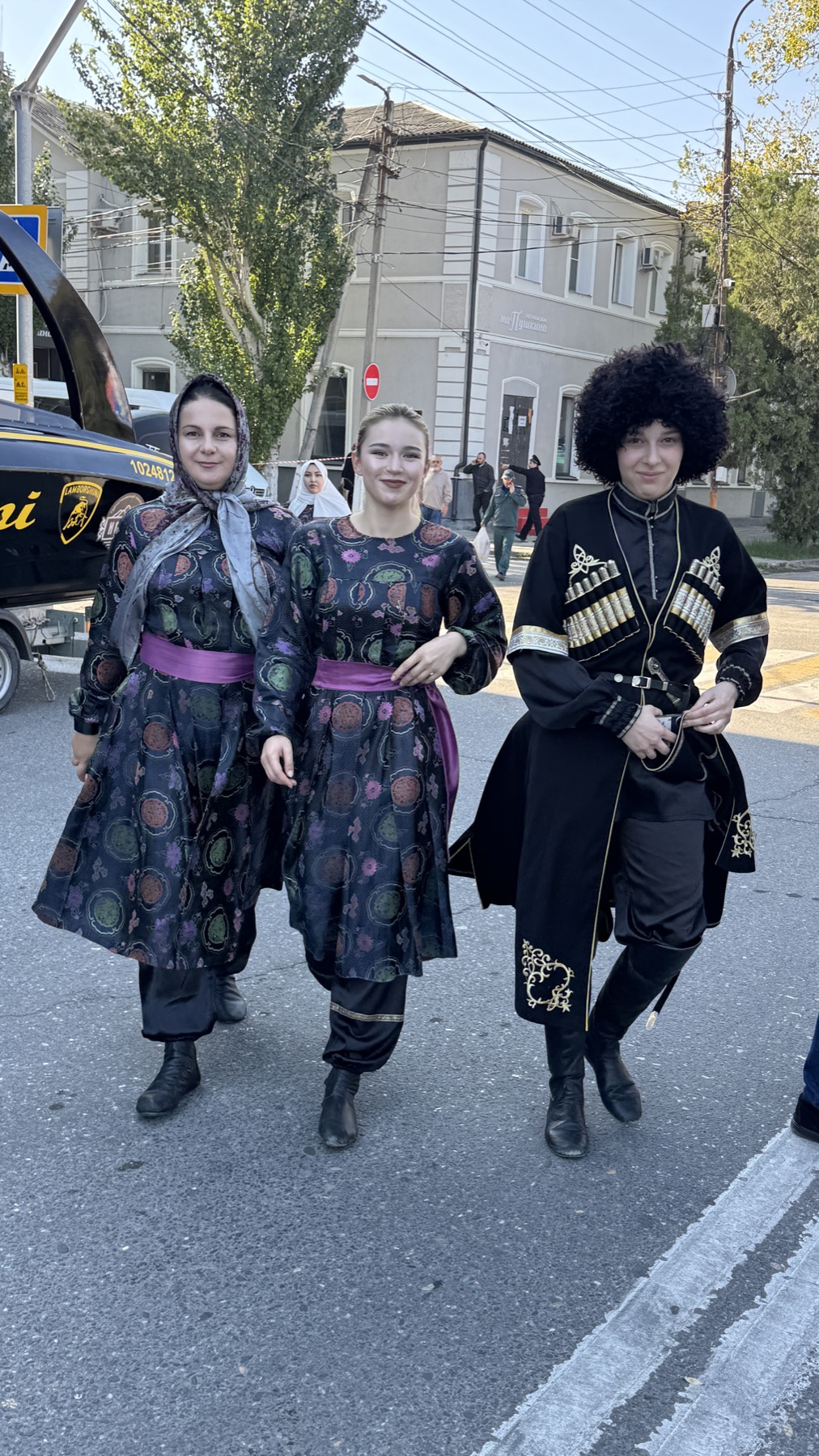 Two women in patterned Dagestani dresses and a man in a black cherkeska with cartridge holders, walking down the street