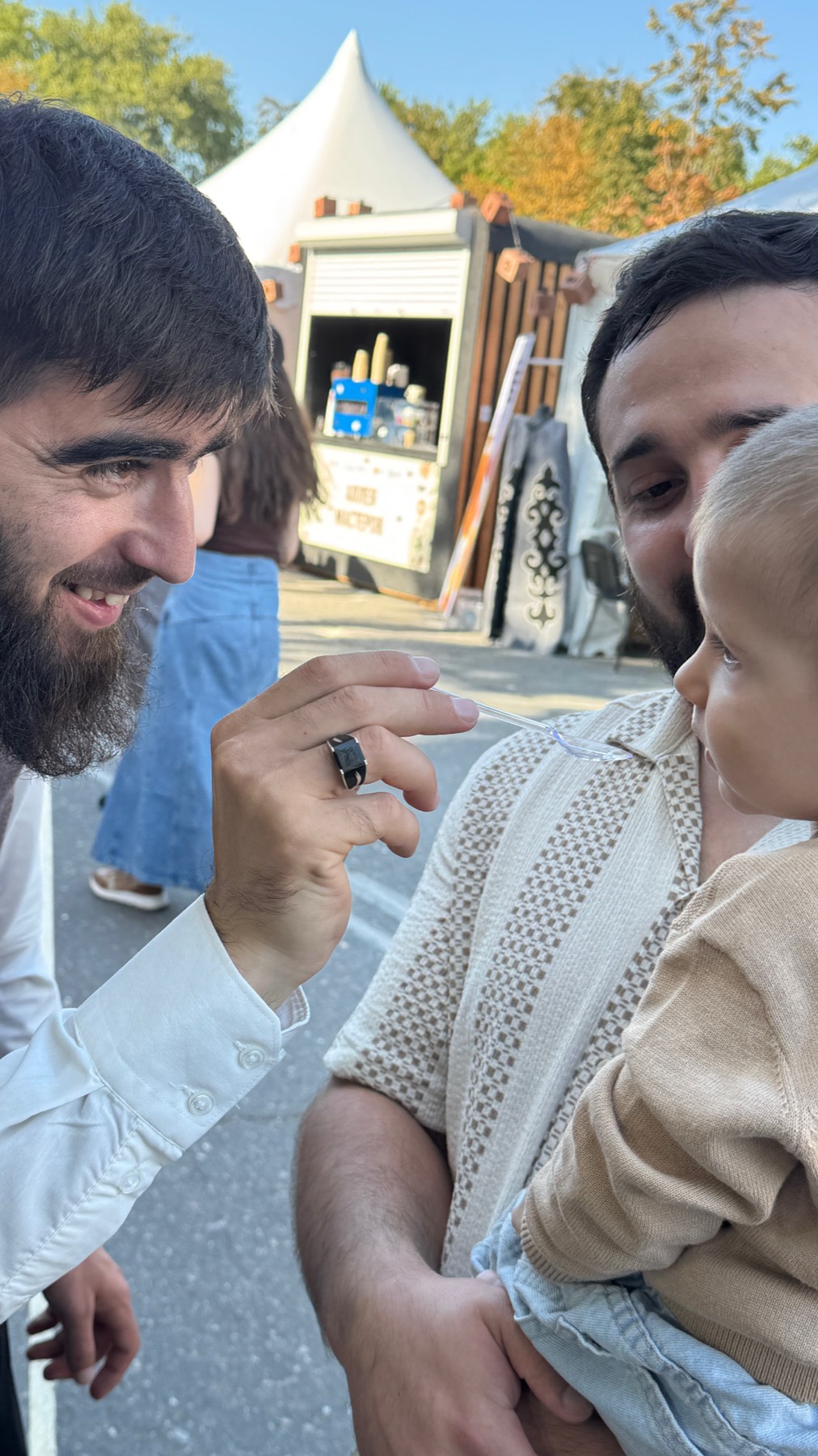A bearded man feeding a small child with a plastic spoon among the festival stands