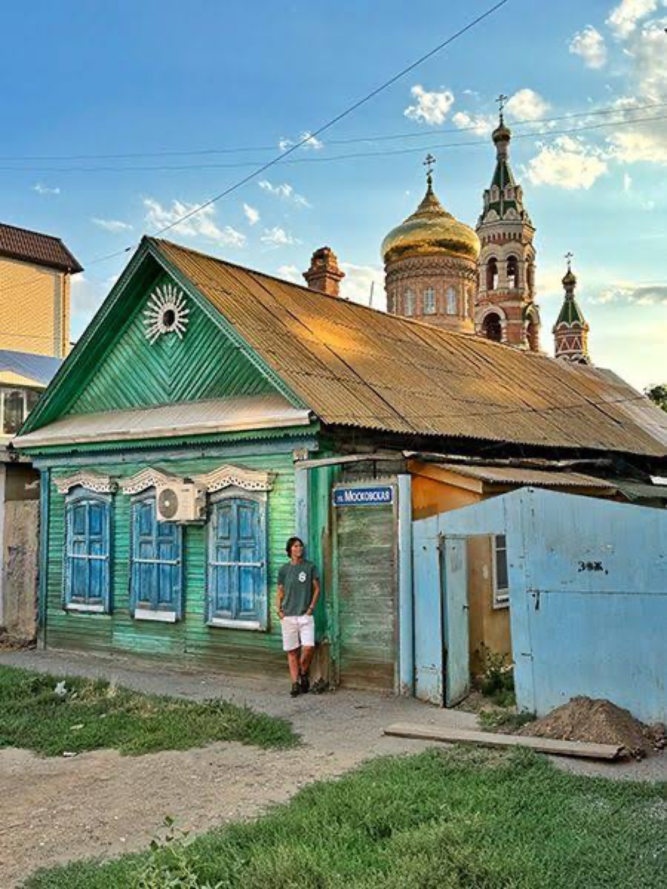 A green wooden house with carved window frames in Astrakhan, with the gold dome of a church behind it