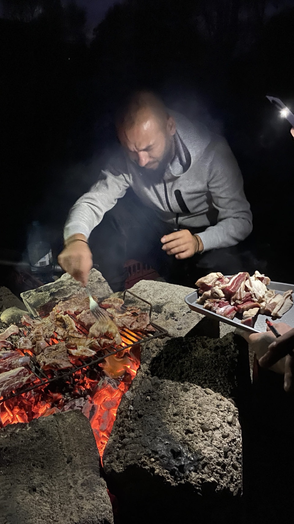 A man grilling meat over an open fire at night